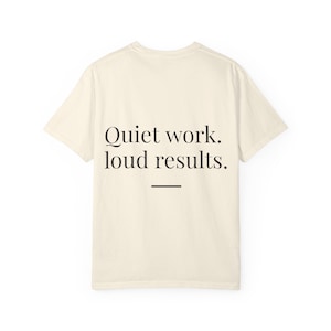 May include: A cream-coloured short-sleeve t-shirt featuring the black printed text "Quiet work. loud results." centred on the back, accompanied by a small horizontal black line positioned directly underneath the text.