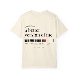 Puede incluir: Una camiseta de manga corta de color crema con texto negro que dice LOADING a better version of me. Debajo del texto hay una barra de progreso con un segmento rojo, seguido del texto 74% - PLEASE BE PATIENT y STILL RISING - N° 08.