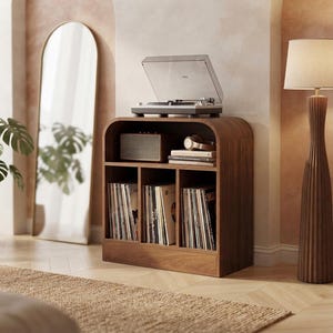 May include: A dark wood record player console with a rounded top edge, featuring an open upper shelf holding a speaker, headphones, and books, and three lower vertical compartments filled with vinyl records, topped with a turntable.