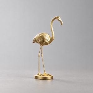 Peut inclure: Une figurine de flamant rose de couleur dorée posée sur une base circulaire. La sculpture présente un corps texturé avec des motifs de plumes détaillés, un long cou courbé et des pattes fines. L'ensemble a une finition métallique brillante.