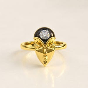 Pode incluir: Um anel de ouro com um design de rosto estilizado e acabamento em ouro amarelo polido. A testa do rosto apresenta uma incrustação circular preta que contém uma única pedra preciosa redonda, transparente e de corte brilhante no centro.