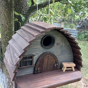 May include: A wooden birdhouse with a curved roof, a dark brown arched door with metal hinges, a small window, and a circular entrance hole. A small wooden bench sits on the front platform. The structure is suspended by white strings.