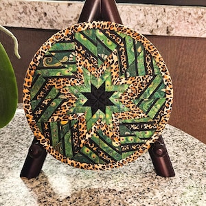May include: A round, handmade coaster with a starburst design. The coaster features a black star in the center, surrounded by green and black geometric shapes, and a leopard print border. The coaster is displayed on a small wooden easel.