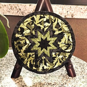May include: A round, quilted coaster with a starburst design. The center star is black, surrounded by olive green and black leaf patterns. The coaster is displayed on a small wooden easel.