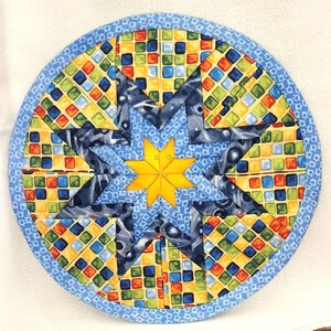 May include: A blue and yellow quilted round pot holder with a star design. The star is made of blue fabric with a yellow center and is surrounded by a border of yellow and blue squares.
