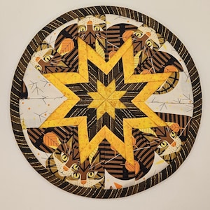 May include: A round, black and gold fabric coaster with a starburst design featuring stylized cats and leaves. The coaster is made of fabric and has a quilted texture.