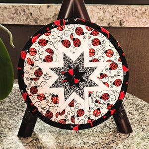 May include: Round quilted coaster featuring a central star design in black and white, surrounded by a white background with red ladybug patterns. The coaster has a black border and is displayed on a small wooden easel.