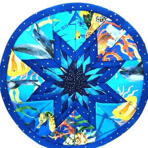 May include: A blue and white fabric coaster with a starburst design featuring sea creatures like dolphins, seahorses, and jellyfish. The coaster has a blue border with white polka dots.