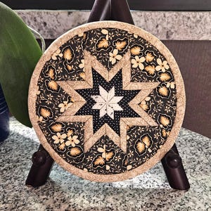 May include: A round, quilted trivet with a star design. The center star is white and black, surrounded by black fabric with white polka dots and floral patterns. The outer edge is a beige floral pattern.