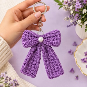 May include: A purple crochet bow keyring featuring a central white pearl bead accent. The bow is attached to a silver-toned metal split ring and a short chain link connector, showcasing a textured yarn construction with two long hanging tails.