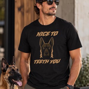Puede incluir: Una camiseta negra de manga corta con un gráfico de la cara de un perro marrón y negro centrado en el pecho. Sobre el perro, el texto dice NICE TO, y debajo del perro, el texto dice TEETH YOU en una fuente negrita de color tostado.