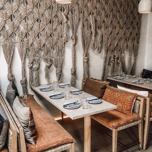 Può includere: Un'area pranzo con un grande arazzo in macramè beige con intricati motivi a nodi. Lo spazio comprende tavoli rettangolari di colore chiaro, sedie in legno con cuscini arancioni e cuscini decorativi fantasia. Due lampade a sospensione bianche.