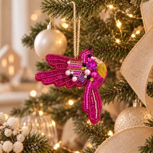Peut inclure: Une décoration en forme de colibri faite à la main, ornée de perles, de paillettes et de cristaux facettés dans des tons fuchsia et rose. L'oiseau présente une aile en paillettes dorées, un grand cristal rose en forme de cœur et une cordelette dorée.