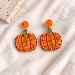 Peut inclure: Une paire de boucles d'oreilles en forme de citrouille ornées de perles orange et rouges. Chaque boucle présente un clou circulaire, une tige en perles dorées métalliques et des strass transparents incrustés sur le corps orange.