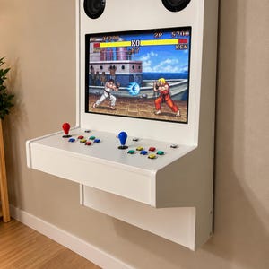 Puede incluir: Una máquina arcade blanca montada en la pared con una pantalla plana que muestra un juego de lucha con los personajes Ryu y Ken. El panel de control incluye dos joysticks con cabezales rojo y azul, además de varios botones de acción.