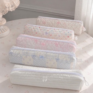 Könnte beinhalten: Fünf rechteckige, gesteppte Stiftemäppchen mit Reißverschluss. Die Designs umfassen ein einfarbig weißes Mäppchen mit kleiner Schleife, ein blau-weißes Blumen-Patchwork-Mäppchen, ein rosa Mäppchen mit Schleifen, ein cremefarbenes mit Herzen und ein cremefarbenes mit Blumen.