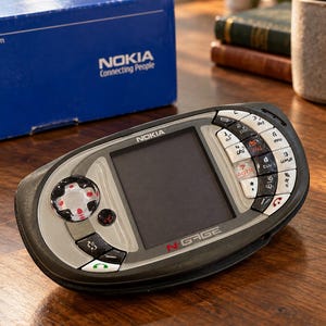 Peut inclure: Un appareil de jeu mobile Nokia N-Gage doté d'un boîtier en plastique gris foncé et argenté, d'un écran central, d'une croix directionnelle à gauche et d'un pavé numérique à droite. L'appareil affiche le logo Nokia et la marque N-Gage sur la face.