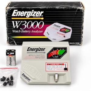 May include: An Energizer W3000 watch battery analyser in a light grey housing with a black analogue gauge, a pulse test switch, and a battery contact point. Includes a 9V battery, a small plastic bag with four black adapters, and the original box.