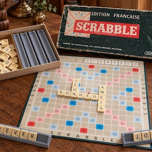 Op de afbeelding: Een vintage Scrabble-bordspelset met een donkergroene doosdeksel met de tekst EDITION FRANCAISE SCRABBLE, een speelbord met lettertegels die JEU, BONHEUR en AMIS spellen, vier grijze letterhouders en een houten bakje met extra lettertegels.