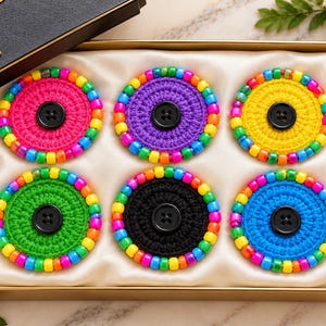 May include: A set of six circular crochet coasters in pink, purple, yellow, green, black, and blue. Each coaster features a border of multicolored plastic beads and a single black four-hole button centered in the middle of the yarn.