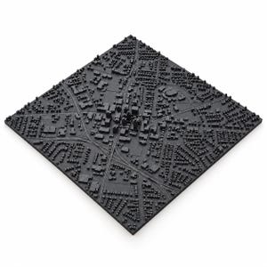 May include: A square, matte black 3D relief map of a city layout, featuring intricate raised details of building blocks, residential neighborhoods, intersecting roads, and a stadium, all presented on a flat, textured base surface.