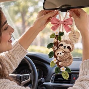 May include: A crochet car hanging ornament featuring a mother sloth and baby sloth clinging to a brown branch with green leaves and white flowers. The ornament includes a pink crochet bow and a heart-shaped wooden tag with the text Good Vibes Every Ride.