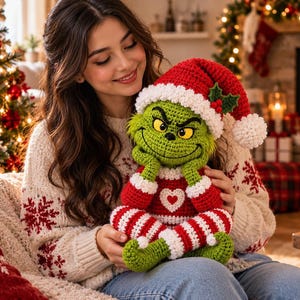 Op de afbeelding: Een gehaakte groene karakterpop met een ondeugende uitdrukking, gekleed in een rood-witte kerstmuts met hulst, een rode trui met een wit hart en een rood-wit gestreepte broek, gemaakt van gedetailleerde gehaakte steken.