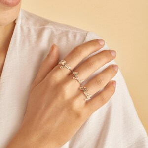 Può includere: Tre anelli color argento indossati sulle dita, ognuno con un nome personalizzato in carattere corsivo. Gli anelli sono sottili fasce metalliche che mostrano i nomi Kylar, Riyah e Shelby con una finitura argentata lucida e riflettente.