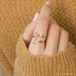 May include: A gold-toned metal ring featuring two stacked, cursive-style name plates. The top name plate displays the name Sophia, and the bottom name plate displays the name Gavin. The ring is worn on a finger, showcasing the script lettering design.