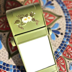May include: An olive green plastic memo pad holder with a hinged top cover. The cover features a hand-painted floral design with white flowers, green leaves, and brown vines. The device includes two side knobs and a blank white paper notepad insert.
