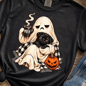 Puede incluir: Una camiseta gris carbón con un gráfico de un fantasma envuelto en una manta de cuadros negros y blancos. El fantasma sostiene una taza negra con el texto MEH y vapor, mientras un carlino negro está sentado junto a una calabaza naranja.