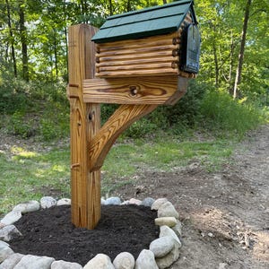 May include: A wooden mailbox designed to resemble a log cabin, featuring a dark green shingled roof and a black metal front door. The mailbox is mounted on a sturdy, vertical wooden post with a diagonal support brace, set in a stone-bordered bed.