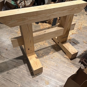 May include: A light brown wooden workbench base constructed from thick, unfinished pine beams. The structure features two vertical support posts connected by a horizontal crossbeam, with two wide feet providing stability for the heavy timber frame.