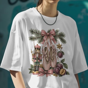 Peut inclure: Un t-shirt blanc avec une impression graphique représentant un soldat casse-noisette, une paire de chaussons de danse roses noués par un grand ruban rose, des branches de sapin, une étoile dorée et des prunes violettes.