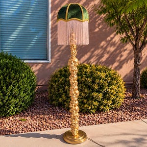 May include: A floor lamp featuring a vertical stack of gold-colored skulls as the base. The lamp is topped with a dark green velvet shade, accented with gold trim and long, light-colored fringe hanging from the bottom edge of the lampshade.