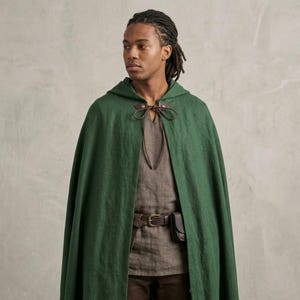Può includere: Un mantello con cappuccio verde bosco caratterizzato da una chiusura con laccetti in pelle marrone sul collo. Il mantello è indossato sopra una tunica marrone con una cintura in pelle marrone che include una piccola tasca. Il capo è testurizzato.