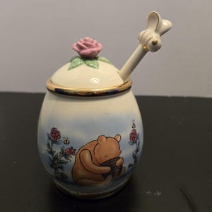 Może przedstawiać: Ceramiczny pojemnik na miód z jasnoniebieskim tłem, ozdobiony ilustracją Kubusia Puchatka, czerwonymi różami i małymi pszczołami. Pokrywka ma uchwyt w kształcie różowej róży oraz pasującą łyżeczkę z pszczołą. Krawędź wykończona złotem.