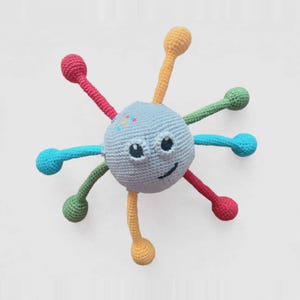 Puede incluir: Un juguete redondo de ganchillo azul claro con una cara sonriente y ojos negros. Presenta ocho brazos largos, delgados y multicolores que se extienden hacia afuera en rojo, amarillo, verde y azul, con extremos pequeños, redondos y bulbosos.