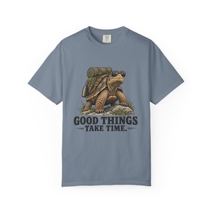 Pode incluir: Uma t-shirt de manga curta azul ardósia com um gráfico de uma tartaruga a usar óculos de sol e uma mochila com um saco de dormir. A tartaruga está sobre uma rocha rodeada de erva. Abaixo do gráfico, o texto diz: GOOD THINGS TAKE TIME.