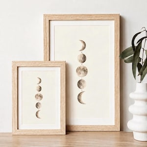May include: Two framed prints featuring a vertical alignment of moon phases in shades of brown and beige. Each print is encased in a light-coloured wooden frame with a white mat border, displaying a series of lunar cycles from crescent to full.