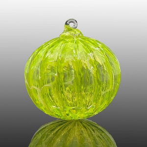 May include: A lime green, spherical glass ornament with a textured surface and a small loop at the top. The ornament is reflected in the surface below, creating a symmetrical image. The glass appears translucent.