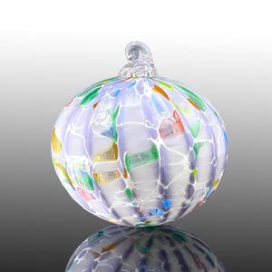 May include: A round, clear glass ornament with a mosaic-like design. The ornament features a variety of colors, including green, orange, blue, and yellow, set against a white and lavender background. A small, clear glass loop is at the top.