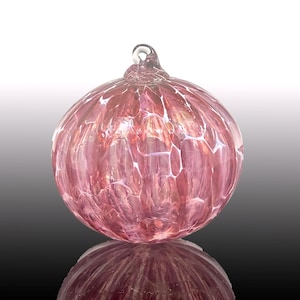 May include: A round, pink glass ornament with a textured surface and a loop at the top for hanging. The ornament has a marbled appearance with white veining throughout. The glass reflects light, creating a shimmering effect.