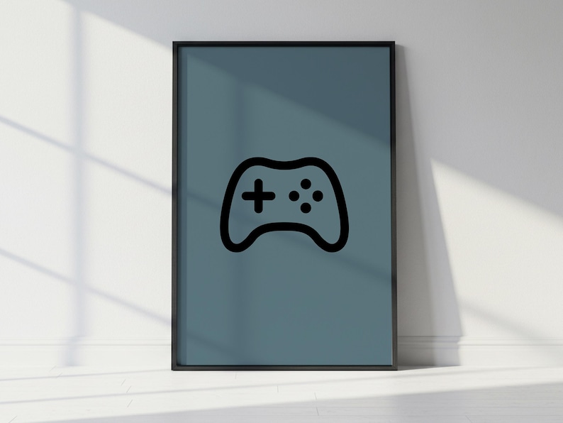 May include: A framed poster featuring a black outline of a video game controller on a teal background. The controller design includes a cross-shaped directional pad and several buttons. The frame is black, and the poster is leaning against a white wall.