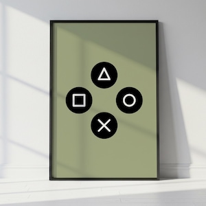 May include: A framed art print featuring a sage green background with four black and white PlayStation controller symbols: a triangle, a square, a circle, and an X. The print is in a black frame and leans against a white wall.