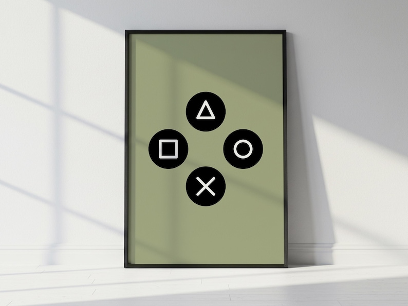May include: A framed art print featuring a sage green background with four black and white PlayStation controller symbols: a triangle, a square, a circle, and an X. The print is in a black frame and leans against a white wall.