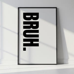 May include: A framed art print with the word "BRUH." in bold, black capital letters against a white background. The print is in a black frame and leans against a white wall, with a shadow cast across the surface.