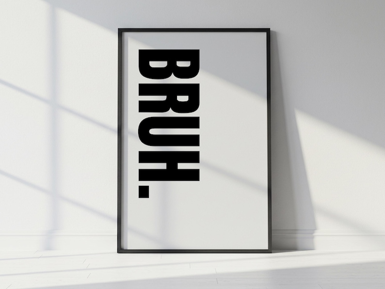 May include: A framed art print with the word "BRUH." in bold, black capital letters against a white background. The print is in a black frame and leans against a white wall, with a shadow cast across the surface.