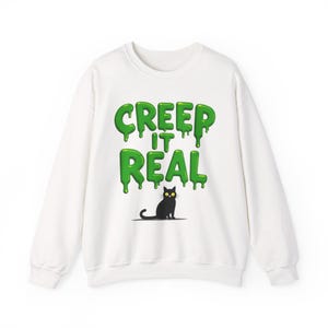 Peut inclure: Un sweat-shirt blanc à manches longues avec le texte vert "CREEP IT REAL" dans une police dégoulinante rappelant de la glu. Sous le texte se trouve une petite illustration de chat noir aux yeux jaunes vifs sur une ligne d'ombre grise.