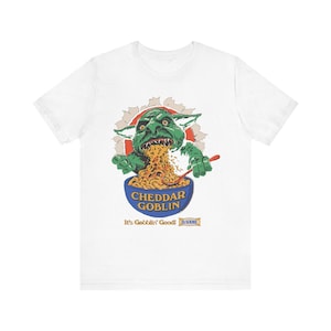 May include: A white short-sleeve t-shirt featuring a graphic of a green goblin eating macaroni and cheese from a blue bowl. The text reads CHEDDAR GOBLIN, It's Gobblin' Good!, and Dr. VANE. The goblin is emerging from a circular, torn-paper effect.